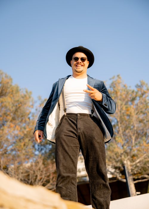 Ein Mann in blauem Jacket, Hut und Sonnenbrille, lachend und auf einen zulaufend, blauer Himmel im Hintergrund